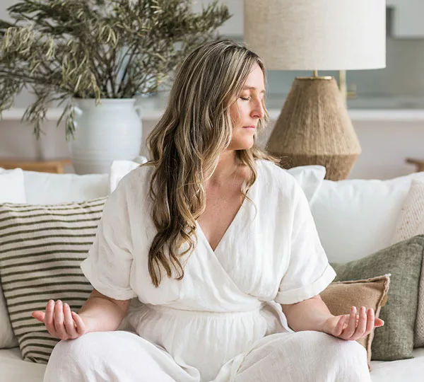 Founder & Director Eleanor Mann, sitting on a linen couch, eyes closed, in a meditation pose
