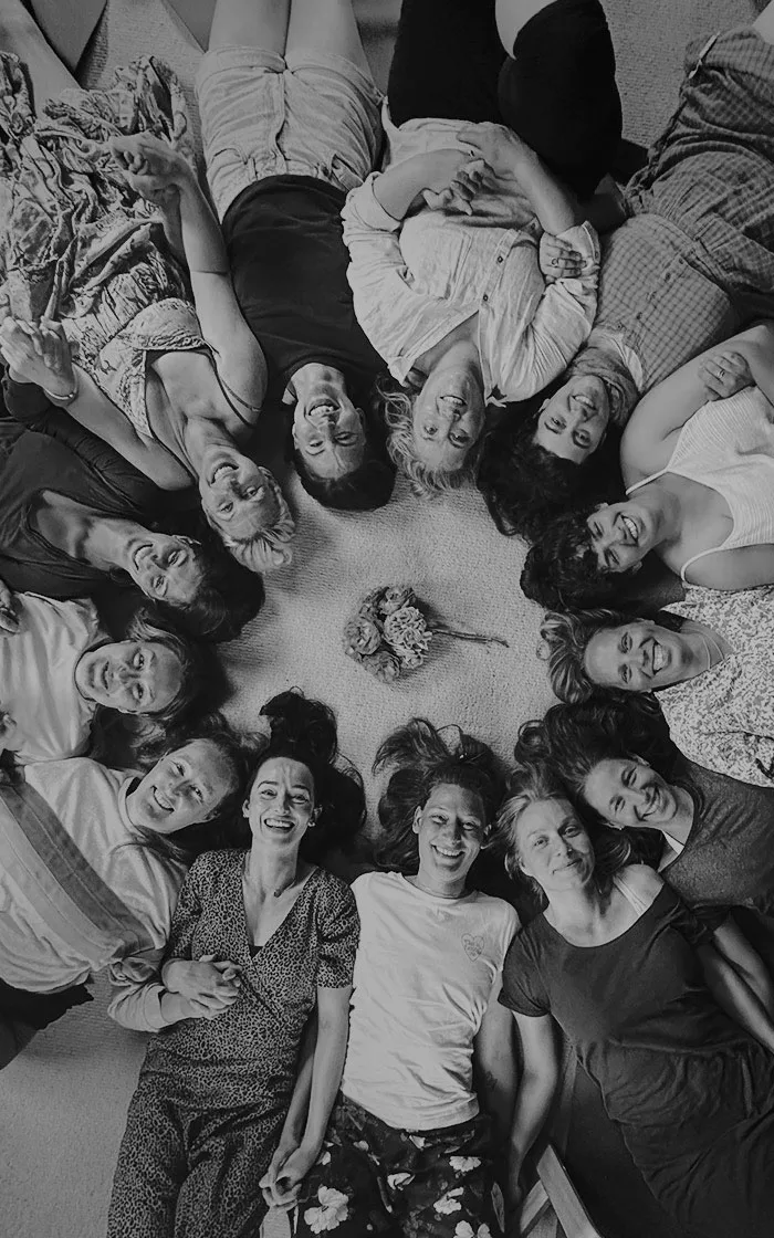 13 Retreat Participants at The School Of Breathwork, lying on their backs in a circle with their heads together and smiling